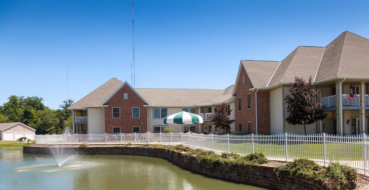 Exterior view of Ancora at Mount Alverna Village showing a multi-story brick and siding building with balconies, a white fence, a pond with a water fountain, and a green and white striped umbrella on the walkway.