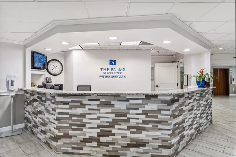 Reception desk area at The Palms of Fort Myers senior living facility with a modern tiled front counter, a wall clock, a mounted TV, a hand sanitizer dispenser, and a potted plant on the counter. The wall behind the desk displays the facility's logo and name.