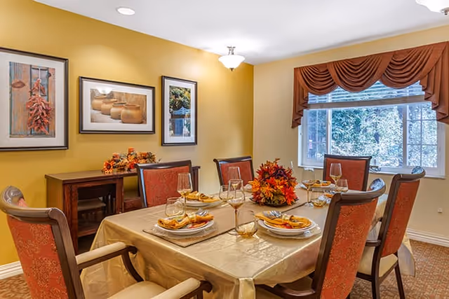 Dining room with a table set for six, upholstered chairs, yellow walls, framed artwork, and a window with a valance.
