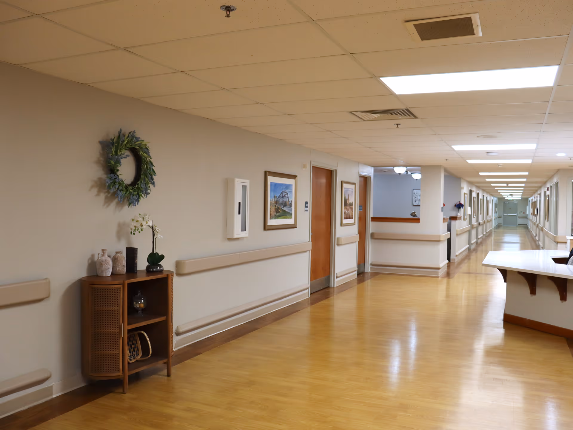A long, well-lit hallway in a senior living facility with wooden floors and beige walls. The hallway features handrails on both sides, framed pictures on the walls, and a small wooden cabinet with decorative items and a wreath hanging above it. There is a reception desk on the right side near the foreground.