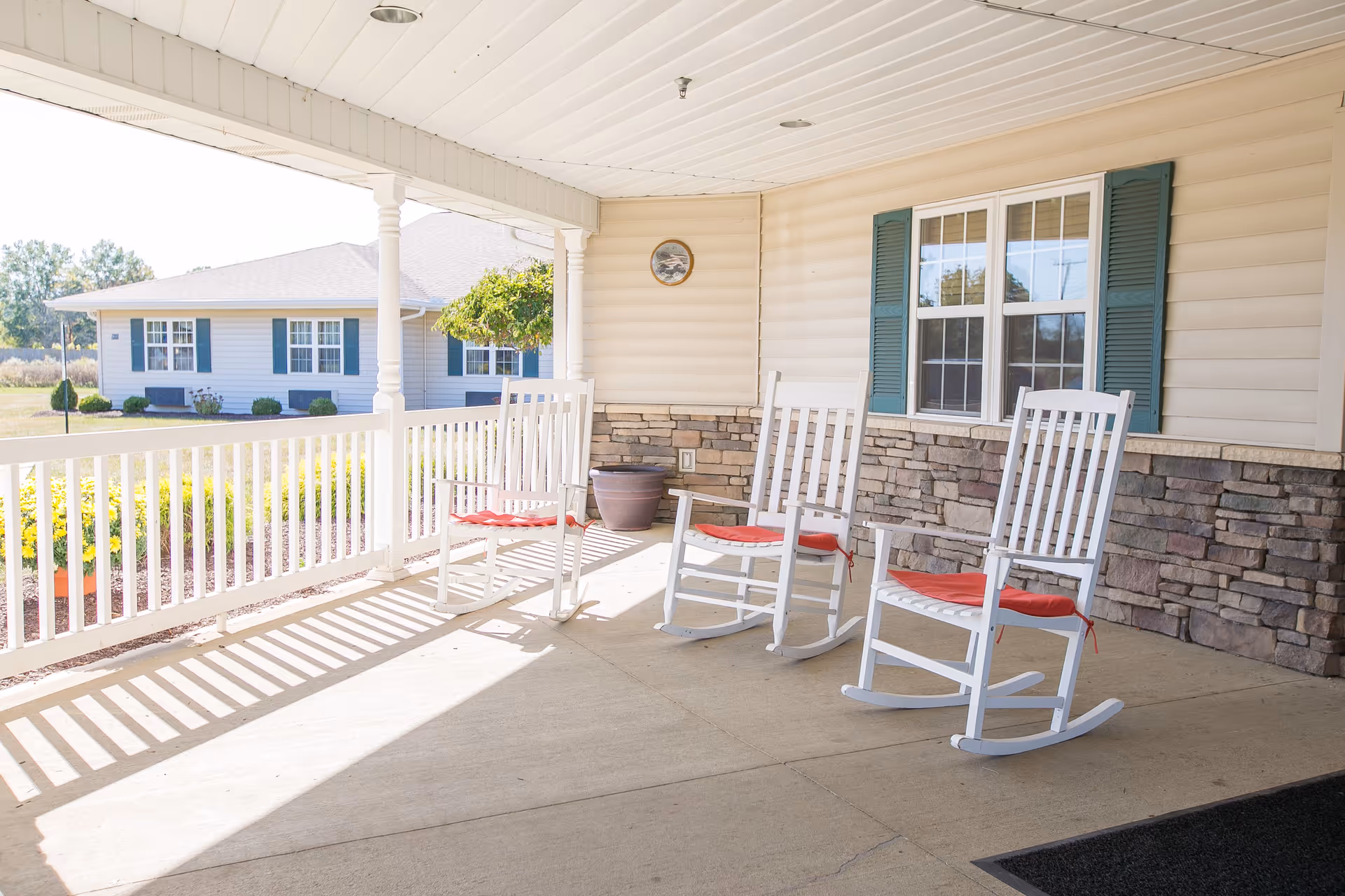 A covered porch area with three white rocking chairs with red cushions, a white railing, and a stone and siding exterior wall. There is a window with green shutters and a potted plant on the porch. In the background, there is a view of another building and some greenery.