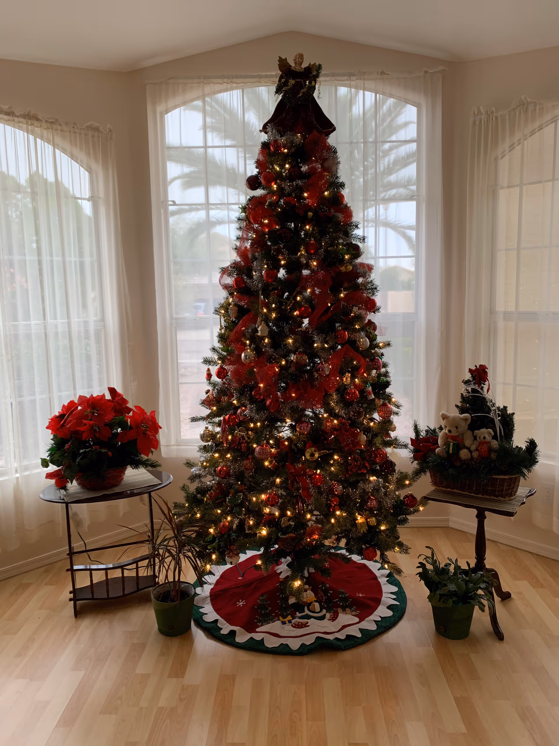 A decorated Christmas tree with lights, red ribbons, and ornaments stands in front of large windows with sheer white curtains. On the left, there is a small table with a red poinsettia plant and a smaller potted plant on the floor. On the right, there is a small round table with a basket containing teddy bears and holiday decorations, and another small potted plant on the floor. The room has light wood flooring.