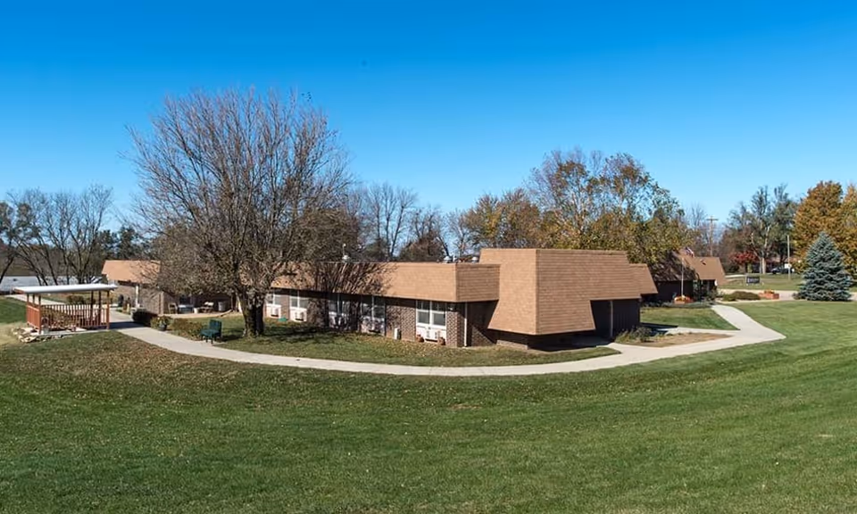 Low single-story brick buildings with brown roofs, a paved walkway, trees, and a large grassy lawn under a clear blue sky.
