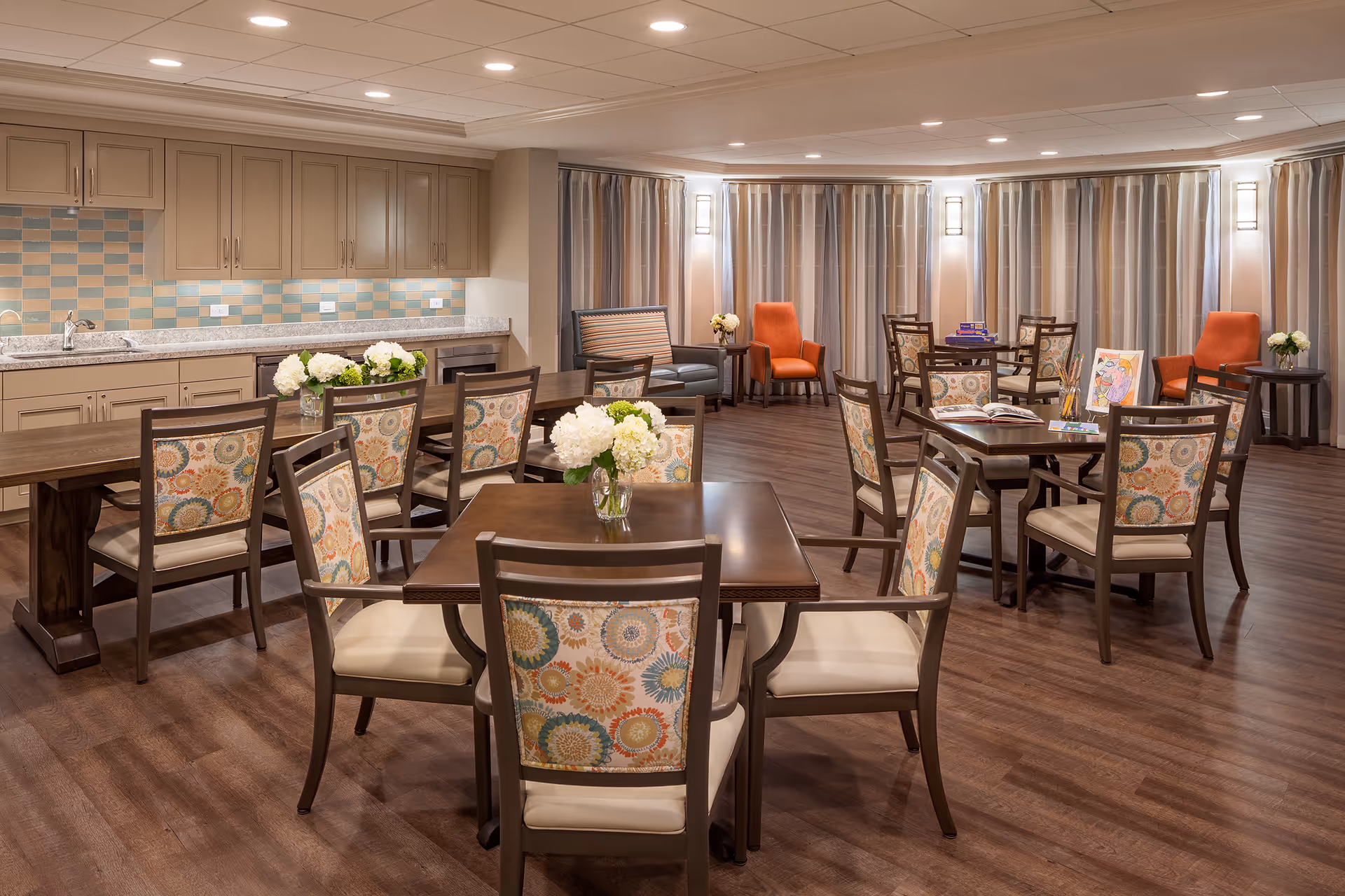 Spacious communal dining and activity room with multiple tables and patterned chairs, a kitchenette along the back wall, and lounge seating.