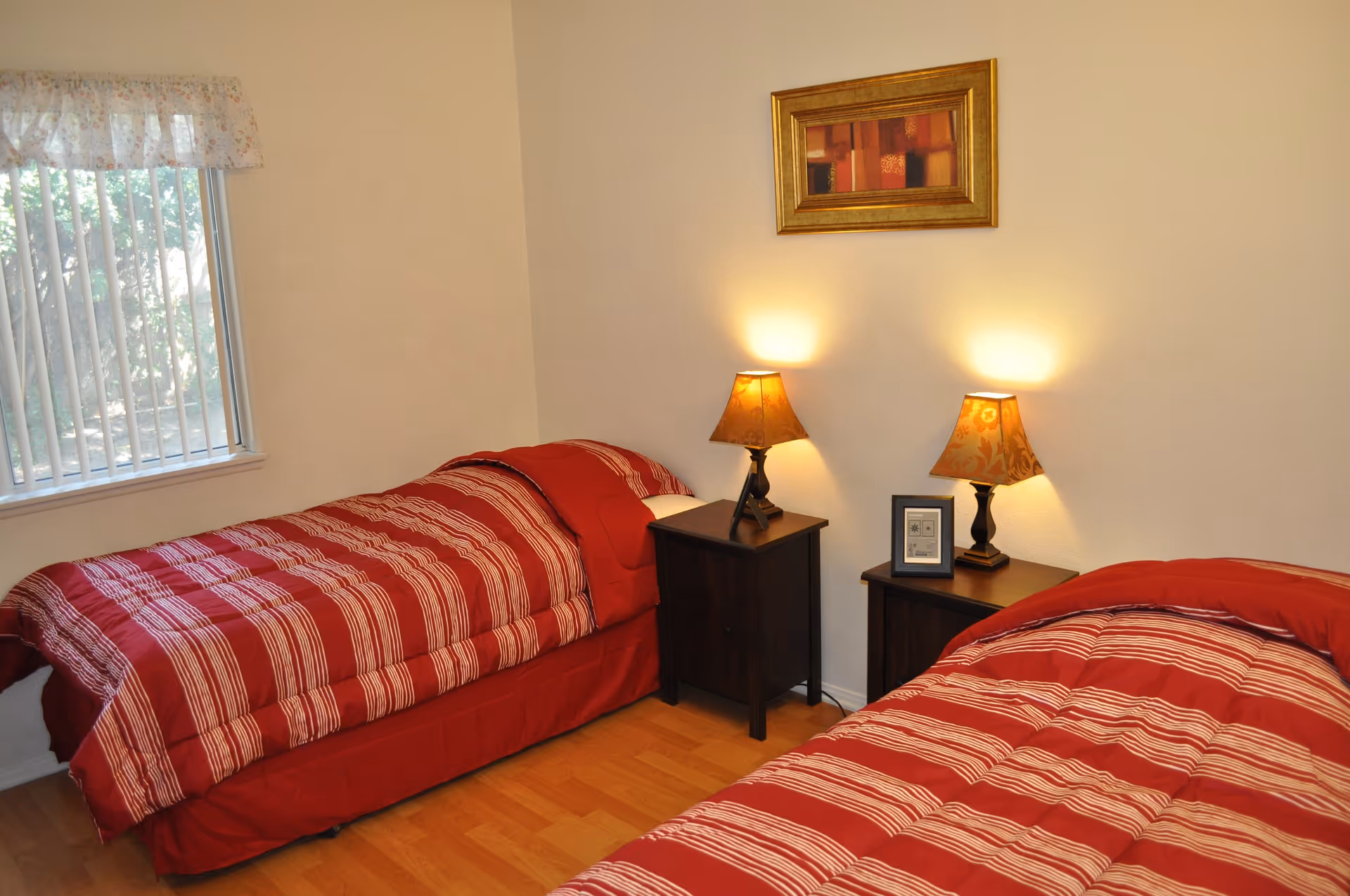 A bedroom with two twin beds covered in red and white striped comforters. Between the beds are two dark wooden nightstands, each with a decorative lamp. A framed picture hangs on the wall above the nightstands. A window with vertical blinds and a floral valance is on the left side of the room, letting in natural light.