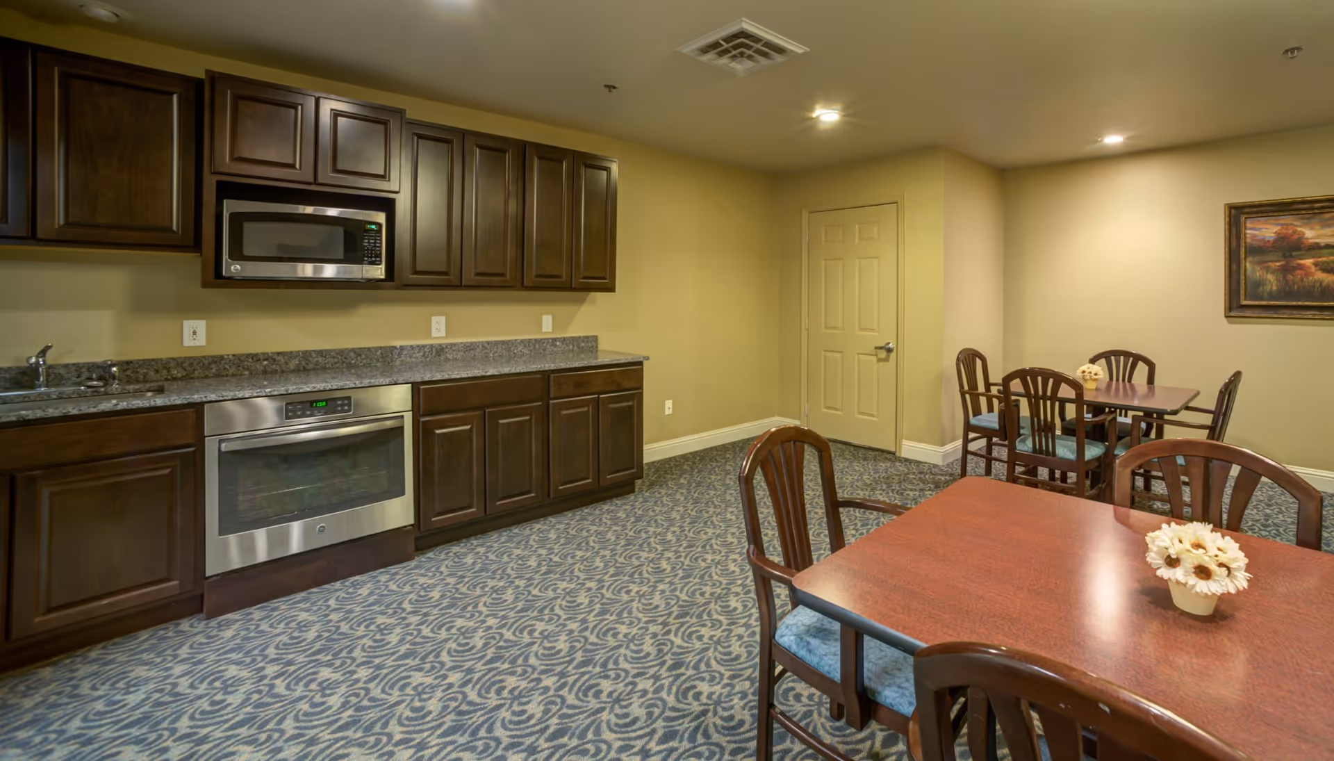 Interior view of a communal kitchen and dining area with dark wood cabinets, a microwave, an oven, and granite countertops on the left side. On the right side, there are two wooden dining tables with chairs, each table decorated with a small flower arrangement. The room has patterned carpet flooring, beige walls, and a framed landscape painting on the wall.