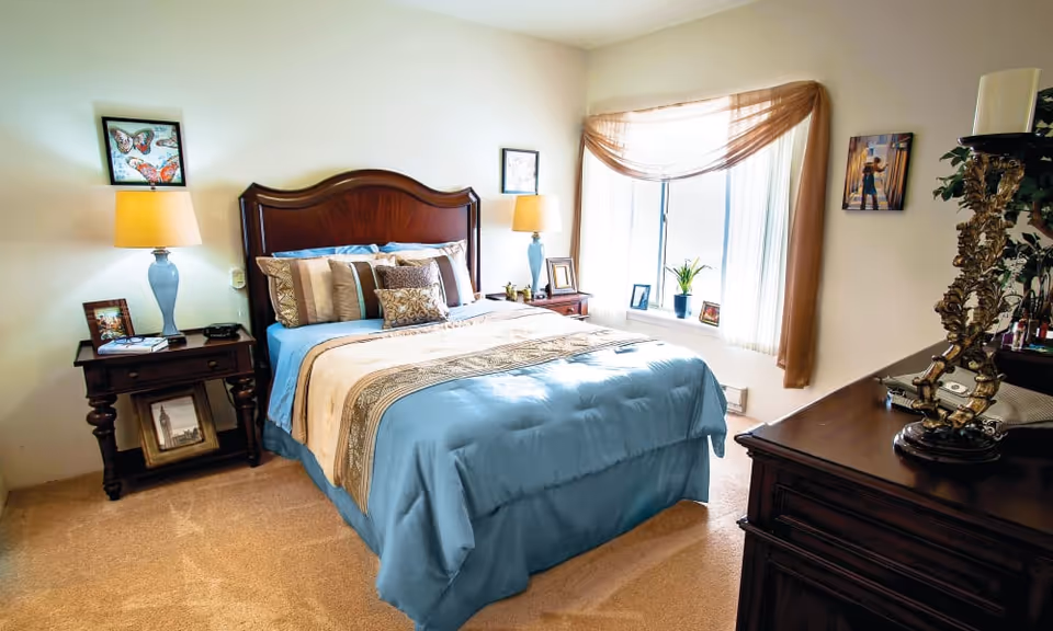 A neatly made bed with blue and beige bedding in a bedroom featuring a wooden headboard. There are two bedside tables with lamps and framed pictures on either side of the bed. A window with sheer curtains and a brown valance lets in natural light. A dark wooden dresser with decorative items is visible on the right side of the room.