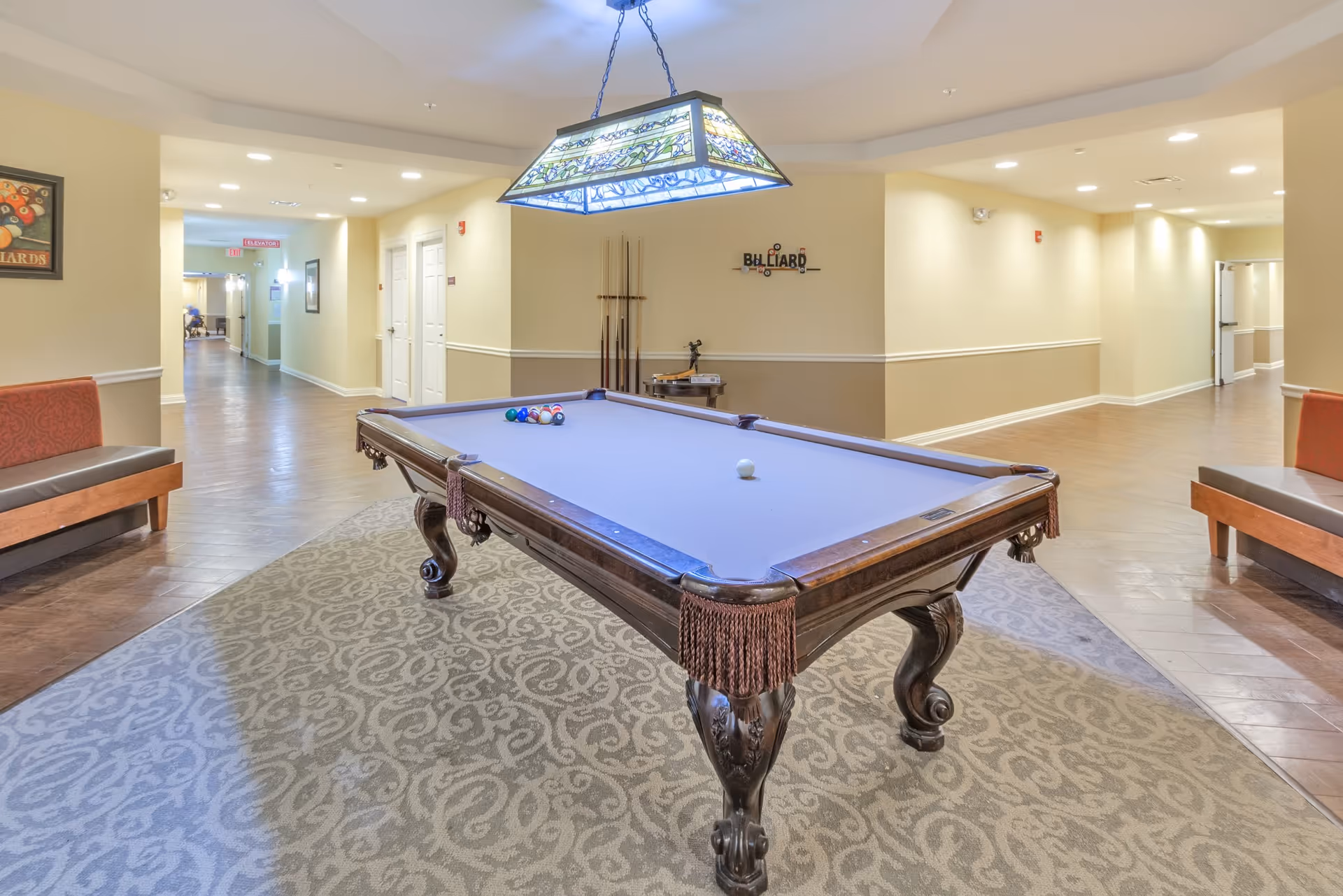 A well-lit interior common area featuring a billiards table with a blue felt surface and a set of billiard balls arranged on it. The table is positioned on a patterned carpet, with two benches on either side. The walls are painted in neutral tones with white trim, and there is a decorative hanging light fixture above the pool table. The hallway extends into the background with doors and framed pictures on the walls.