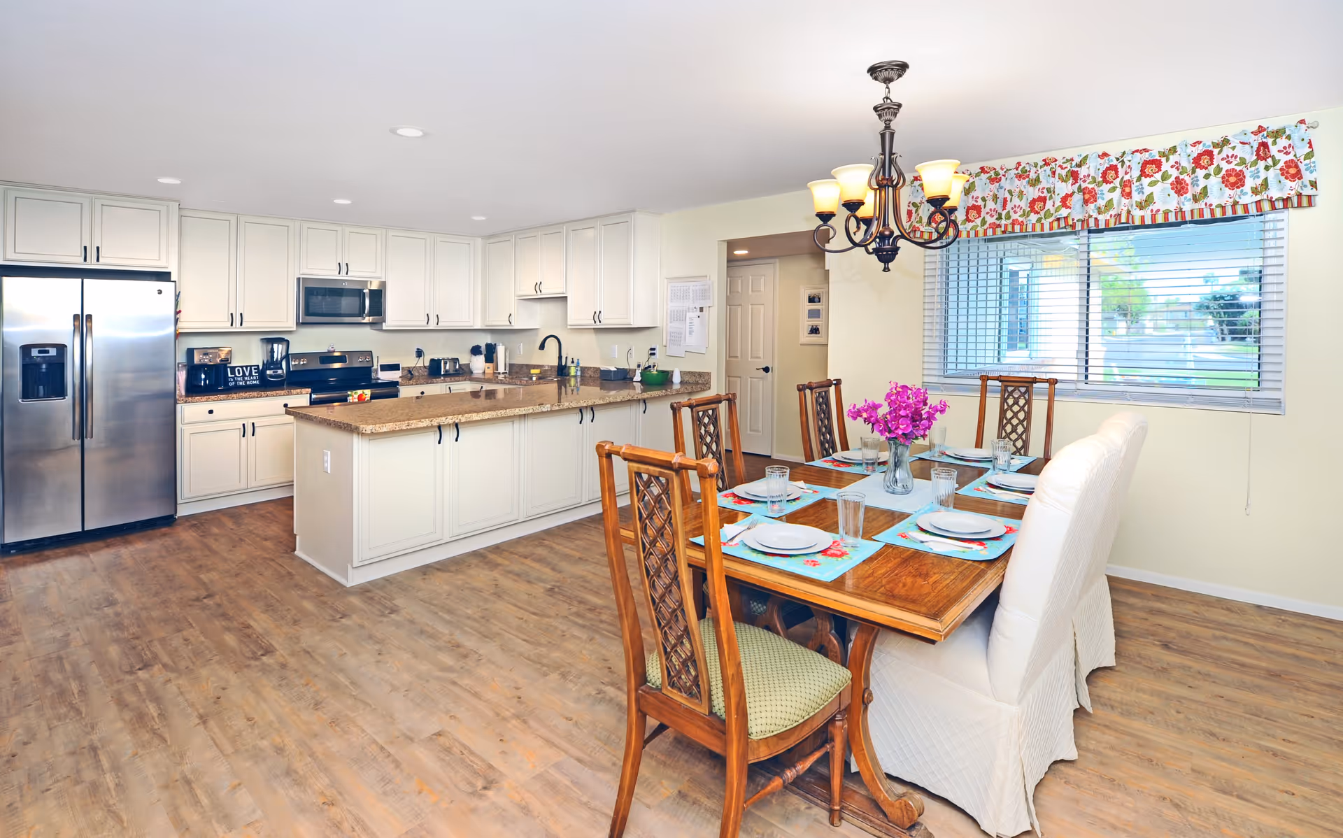 Bright and spacious kitchen and dining area with white cabinets, stainless steel refrigerator and appliances, a large island with granite countertop, and a wooden dining table set for six with colorful placemats and a vase of purple flowers. A window with floral valance lets in natural light.