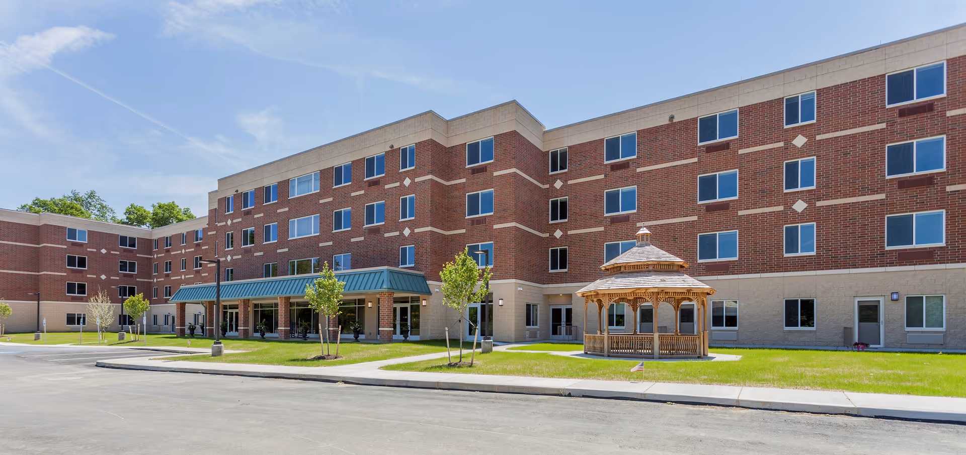 Exterior view of a multi-story senior living facility building with red brick and beige accents. The building has many windows and a green metal awning over the entrance. In front of the building, there is a well-maintained lawn with small trees and a wooden gazebo. The sky is clear and blue.
