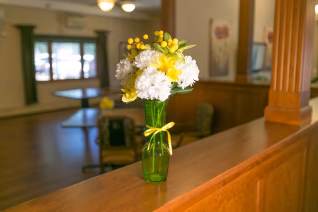 A green glass vase with a yellow ribbon tied around it, holding a bouquet of white and yellow flowers, placed on a wooden ledge in a room with tables and chairs in the background.