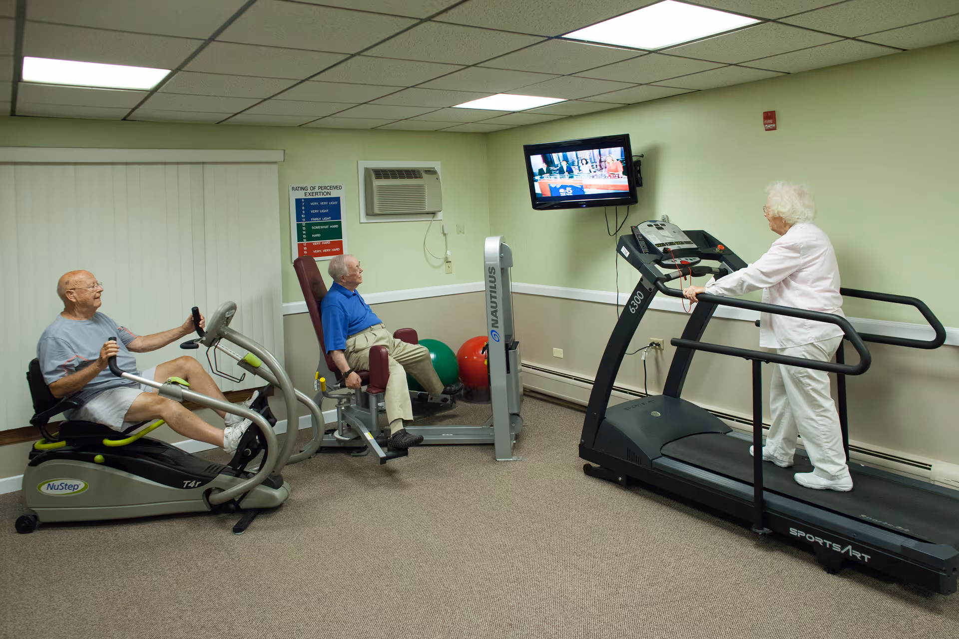 Three elderly individuals exercising in a fitness room. One man is using a recumbent exercise bike, another man is seated on a leg exercise machine, and a woman is walking on a treadmill while watching a wall-mounted television. The room has light green walls, a beige carpet, and fluorescent ceiling lights.