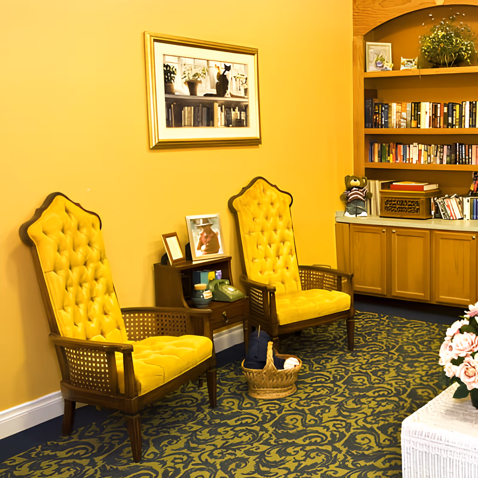 A cozy sitting area with two vintage yellow tufted armchairs placed against a yellow wall. Between the chairs is a small wooden side table with framed photos and a green rotary phone. A basket with knitting supplies sits on the patterned carpet in front of the chairs. To the right, there is a built-in wooden bookshelf filled with books and decorative items, including a teddy bear. A framed picture of a cat on a windowsill hangs on the wall above the chairs.
