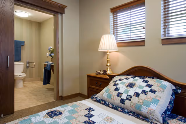 Cozy bedroom with a quilted bed, wooden nightstand and lamp, and an en-suite bathroom visible through an open doorway.
