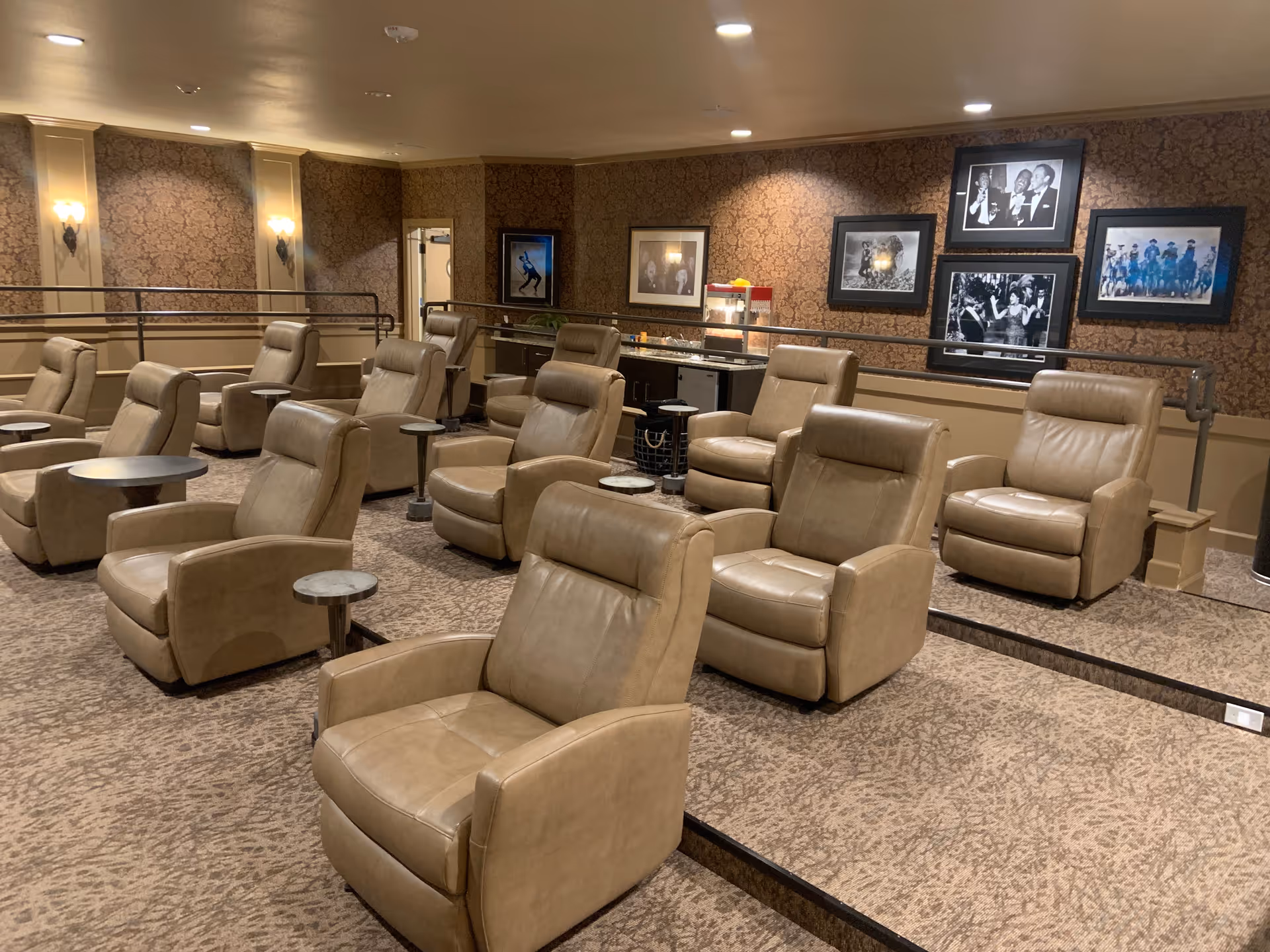 A cozy theater-style room with multiple rows of beige leather recliner chairs, each paired with a small round side table. The room has patterned carpet and wallpaper with framed black and white photos on the walls. There is a popcorn machine and a counter with snacks at the back of the room.