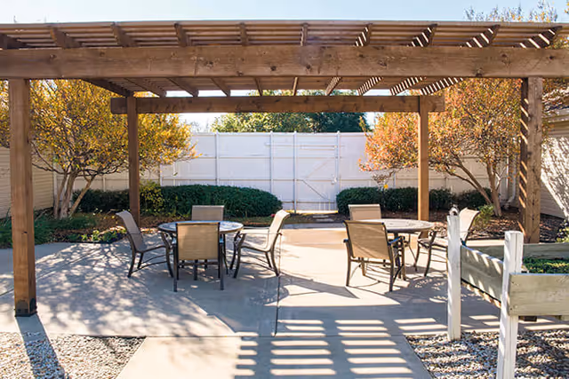 Outdoor patio area with two round tables and several chairs under a wooden pergola. The area is surrounded by small trees and bushes, with a white fence in the background.