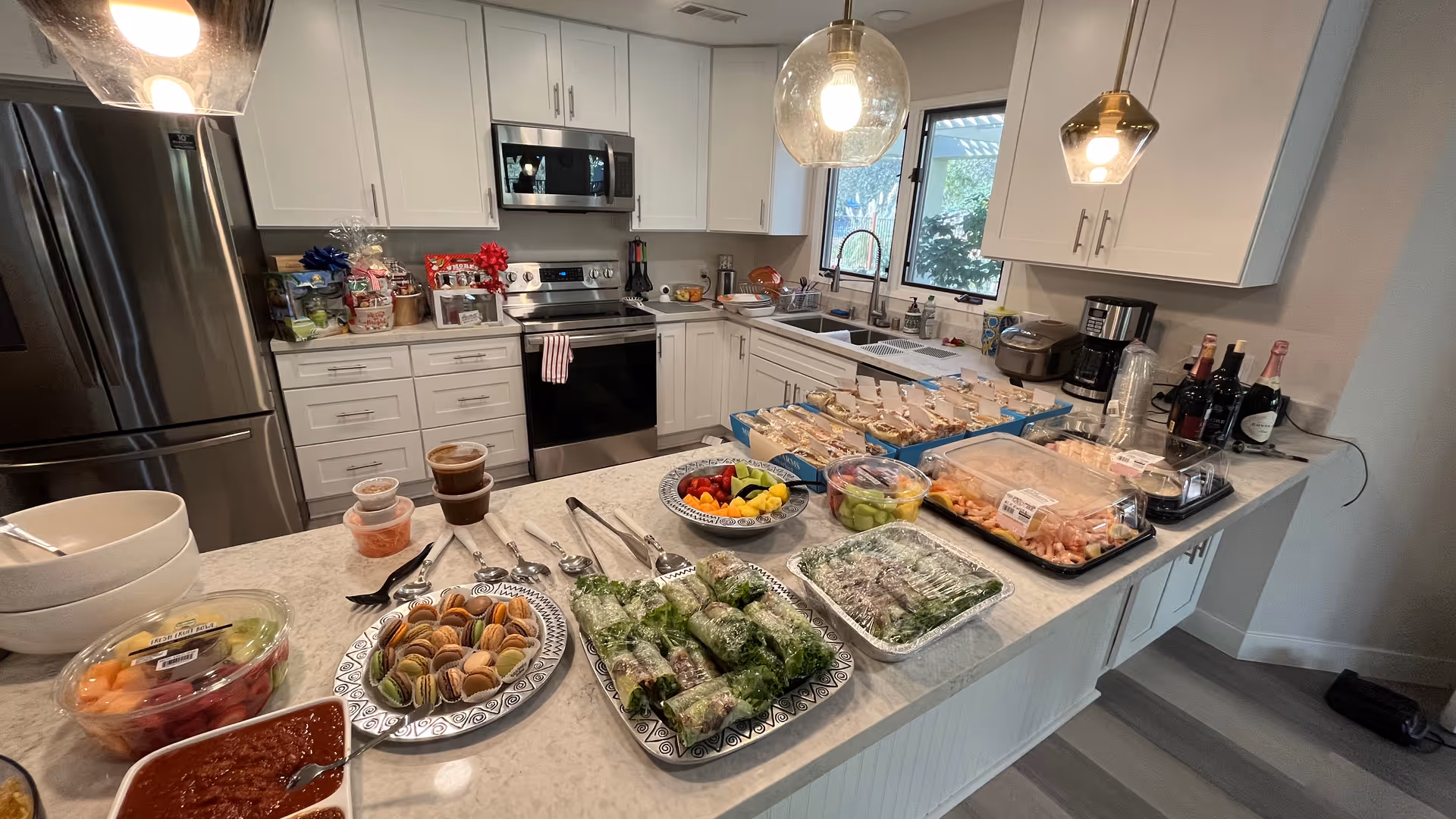 A modern kitchen with white cabinets and stainless steel appliances. The countertop is filled with various food items including sandwiches, spring rolls, fruit platters, macarons, shrimp, and sauces. Two pendant lights hang above the counter, and a window above the sink shows greenery outside.