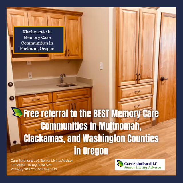 A kitchenette area in a memory care community featuring wooden cabinets, a countertop with a small sink, and a wooden door. The floor is wooden, and the space is clean and well-lit.