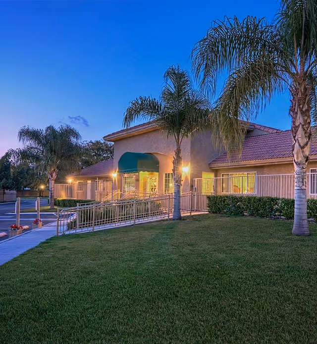 Exterior view of Desert Hills Memory Care Center at dusk, showing a single-story building with a green awning over the entrance, palm trees, well-maintained grass, and outdoor lighting.