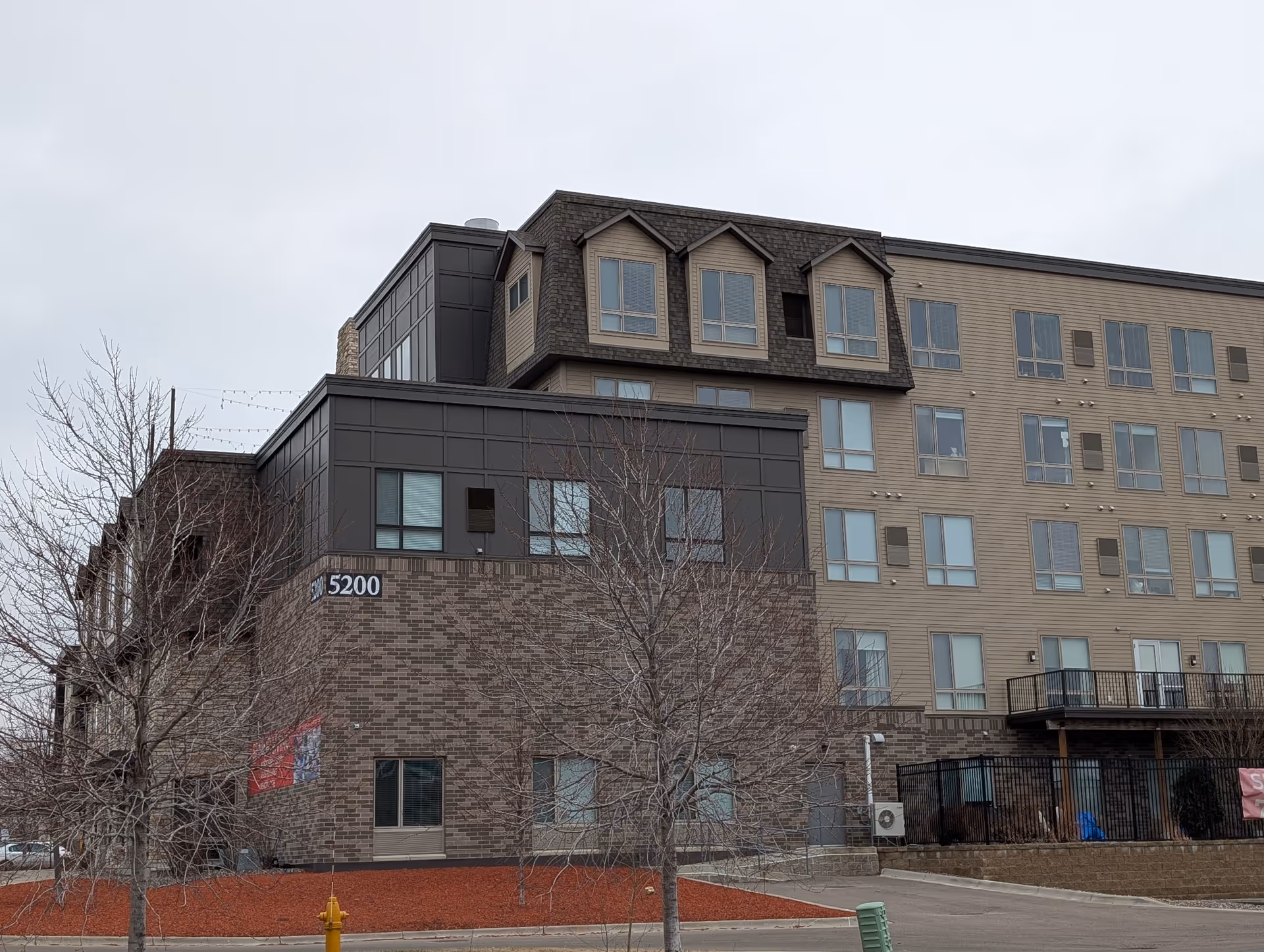 Exterior view of a multi-story senior living facility building with a combination of brick and siding exterior walls, multiple windows, and a few leafless trees in the foreground under an overcast sky.