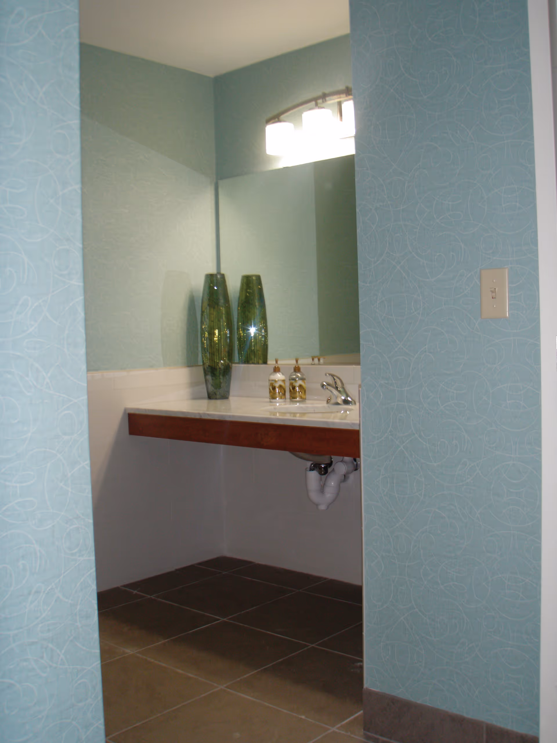 A bathroom vanity area with a large mirror, two green decorative vases, a sink with a faucet, and two soap dispensers. The walls are painted light blue with a subtle swirl pattern, and the floor is tiled in brown.