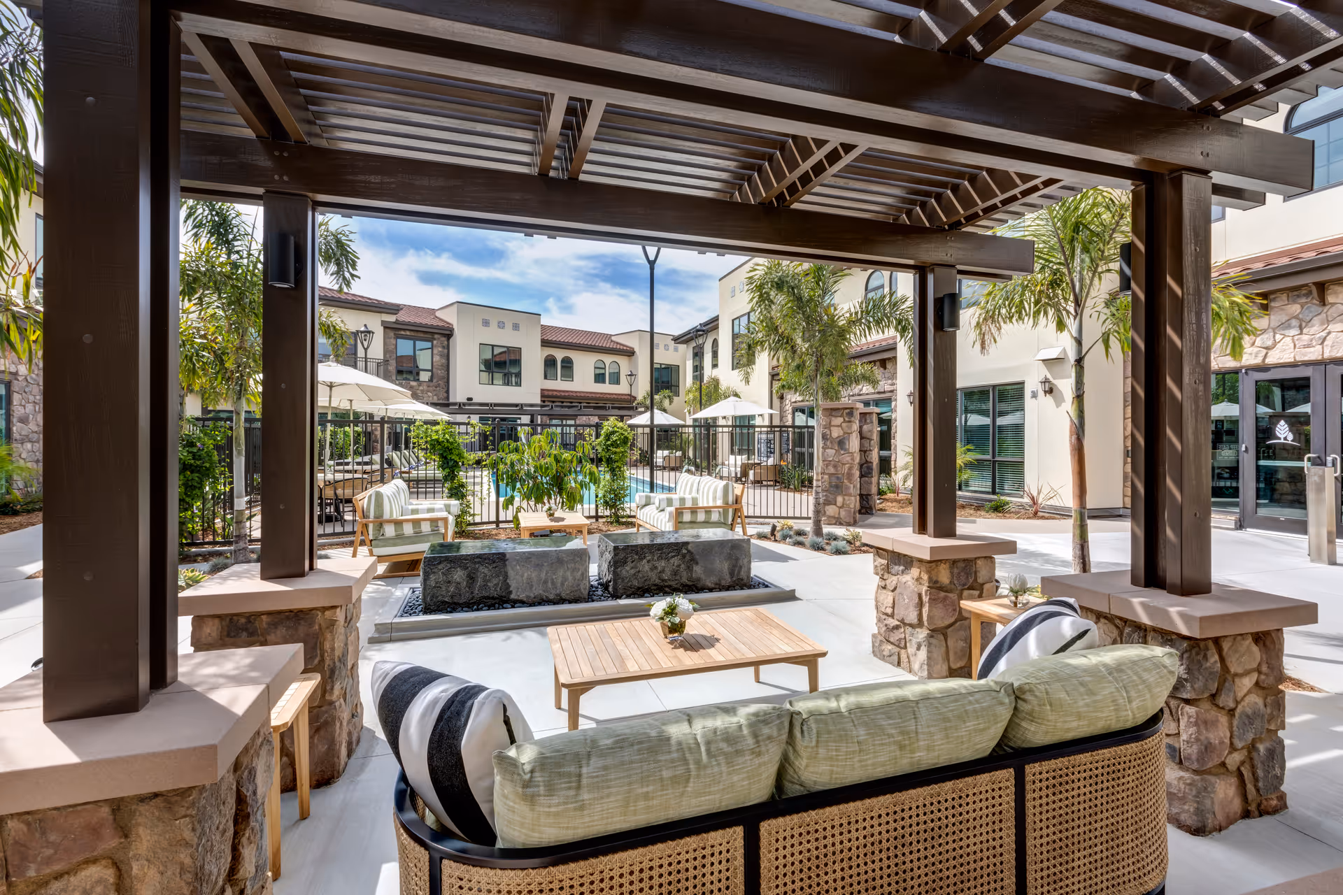 Shaded outdoor courtyard with cushioned seating under a pergola, a central water feature, and views of the building and pool area.