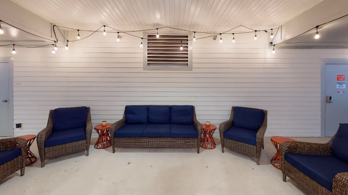 An indoor seating area with wicker furniture featuring navy blue cushions, including a three-seat sofa and four armchairs. There are small red side tables next to the chairs and sofa. String lights with exposed bulbs are hung across the white paneled wall above the seating. Two gray doors are visible on either side of the wall.
