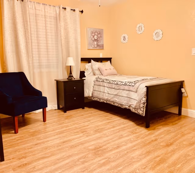 A cozy bedroom with a single bed featuring patterned bedding and multiple pillows. Next to the bed is a black nightstand with a lamp. A dark blue upholstered chair is positioned near a window with white curtains. The walls are painted a warm beige color and decorated with three small white-framed pictures. The floor is light wood.