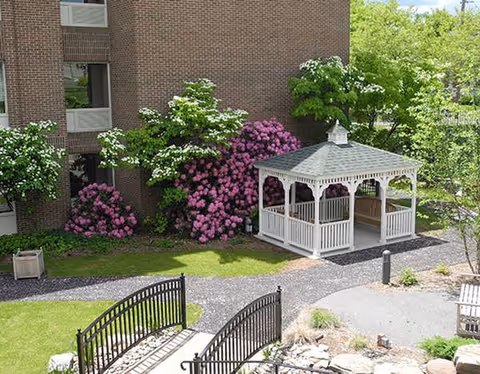 Outdoor courtyard with a white gazebo, blooming shrubs, and brick building.