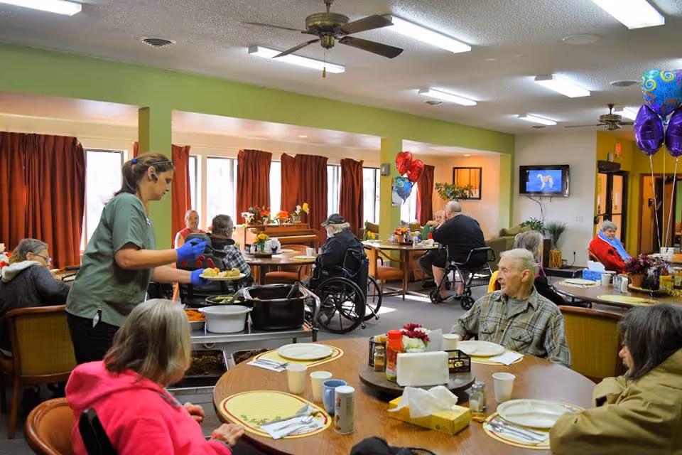 A dining room in a senior living facility with several elderly residents seated at round tables. A staff member wearing gloves and a headset is serving food from a cart. The room has green walls, large windows with red curtains, ceiling fans, and balloons in the background. A television is mounted on the wall.