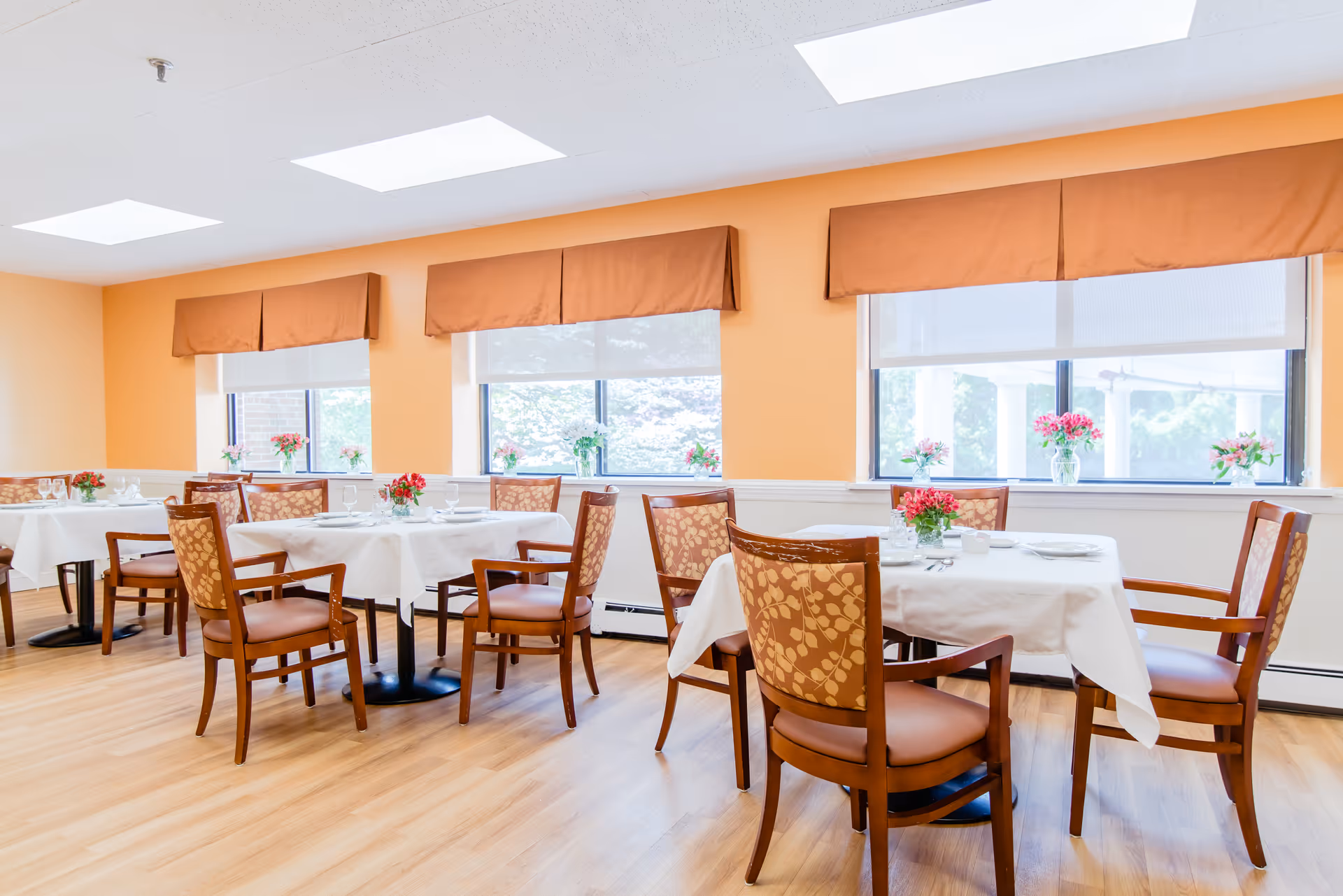 A bright dining room with several tables covered in white tablecloths, each set with plates, glasses, and silverware. Wooden chairs with patterned upholstery surround the tables. The room has large windows with orange valances and small vases of pink flowers on the windowsills and tables. The floor is light wood, and the walls are painted a warm peach color.