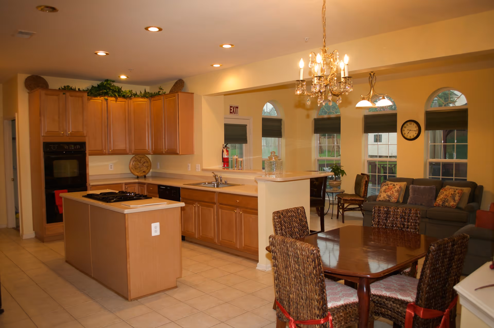 A warm, inviting kitchen and dining area with wooden cabinets, a kitchen island with a stovetop, a double sink, and a dining table with four wicker chairs tied with red ribbons. The room is well-lit with recessed ceiling lights and a chandelier above the dining table. In the background, there is a living area with a sofa, chairs, a clock on the wall, and several arched windows with green blinds.