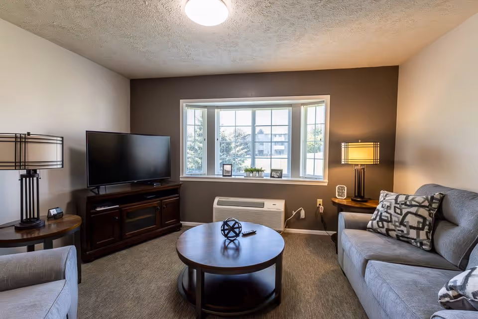 A cozy living room with a large window featuring blinds and a view of trees and buildings outside. The room has a gray couch with patterned pillows, a round wooden coffee table with a decorative object and remote control, a flat-screen TV on a dark wooden stand, and two matching table lamps on either side of the room.