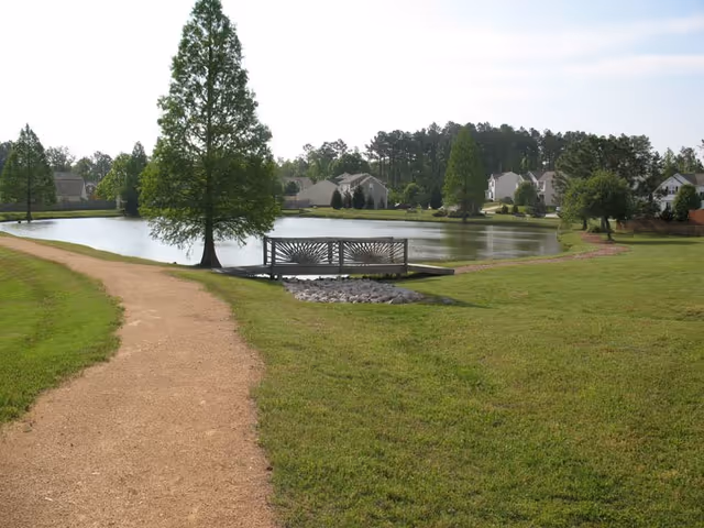 Grassy park area with a gravel path, a small decorative bridge over a pond, and trees.