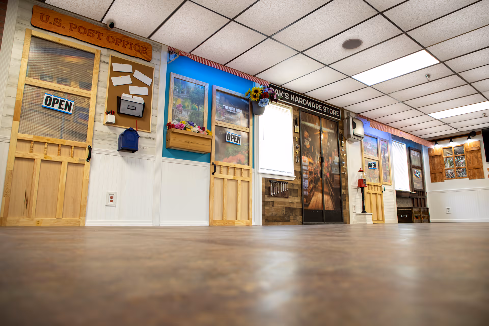 Indoor hallway area designed to look like a small town street with storefront doors labeled U.S. Post Office, Oak's Hardware Store, and other shops. The walls are decorated with signs, windows, flower boxes, and bulletin boards, creating a quaint and inviting atmosphere.