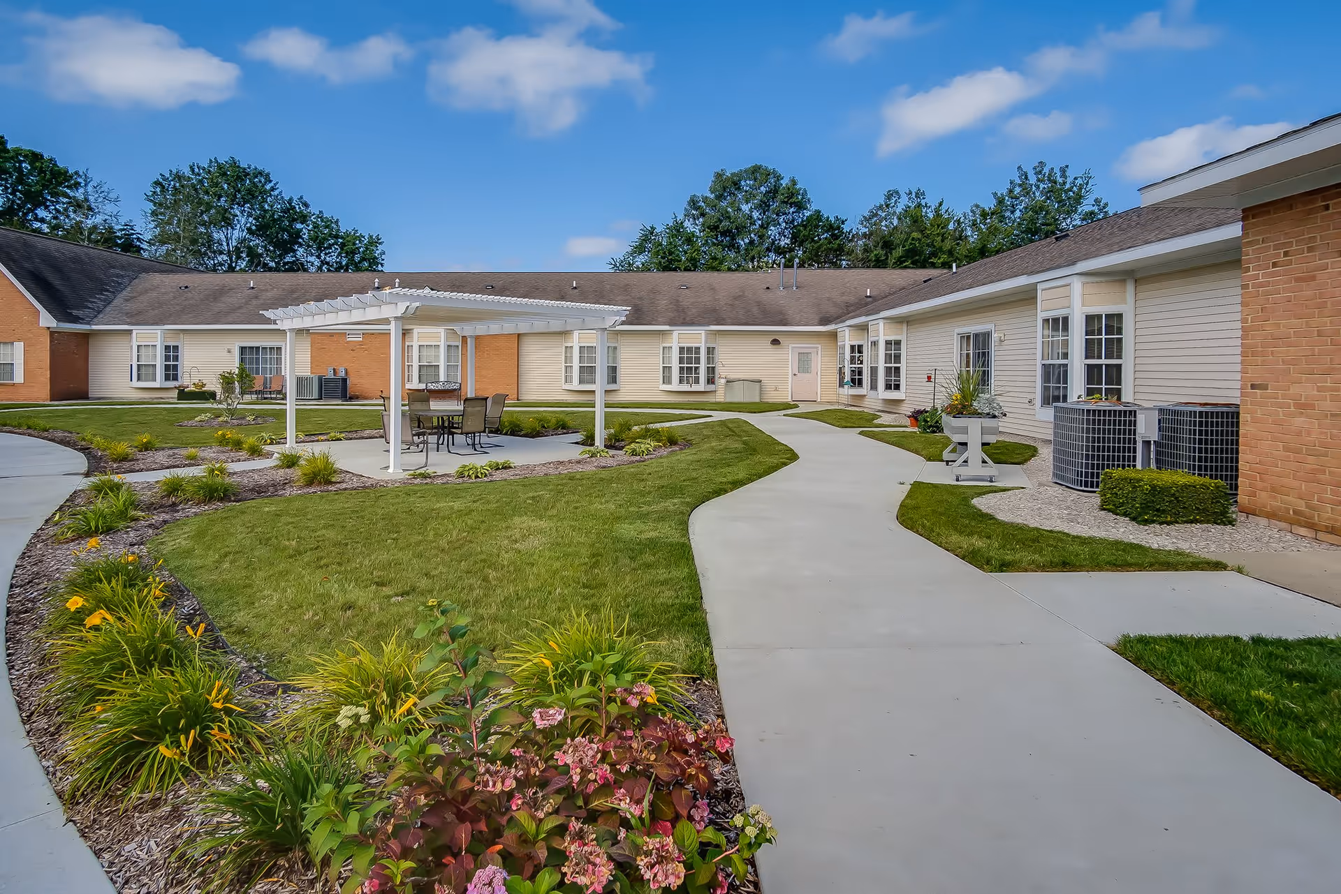 Outdoor courtyard area of a retirement village with a paved walkway, green grass, flower beds, and a white pergola covering a table with chairs. The building surrounding the courtyard has beige siding and brick accents with multiple windows and doors.