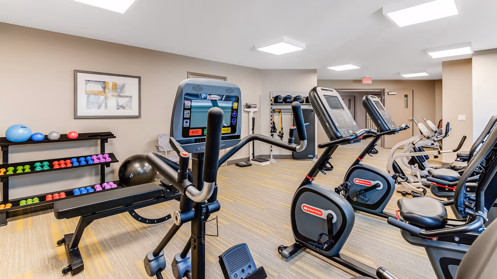 A fitness room with exercise equipment including stationary bikes, a weight bench, dumbbells in various colors on racks, exercise balls, and resistance bands hanging on the wall. The room has beige walls, carpeted flooring, and bright ceiling lights.