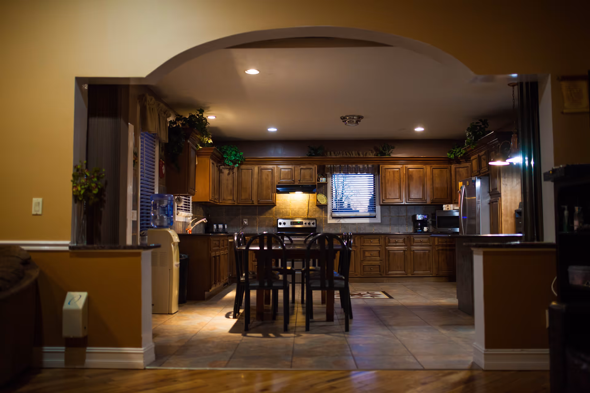 Interior view of a kitchen with wooden cabinets, a dining table with four chairs in the center, a stainless steel stove and refrigerator, a window with blinds above the sink, and various plants placed on top of the cabinets. The kitchen is seen through an arched opening from an adjacent room with warm yellow walls and tiled flooring.