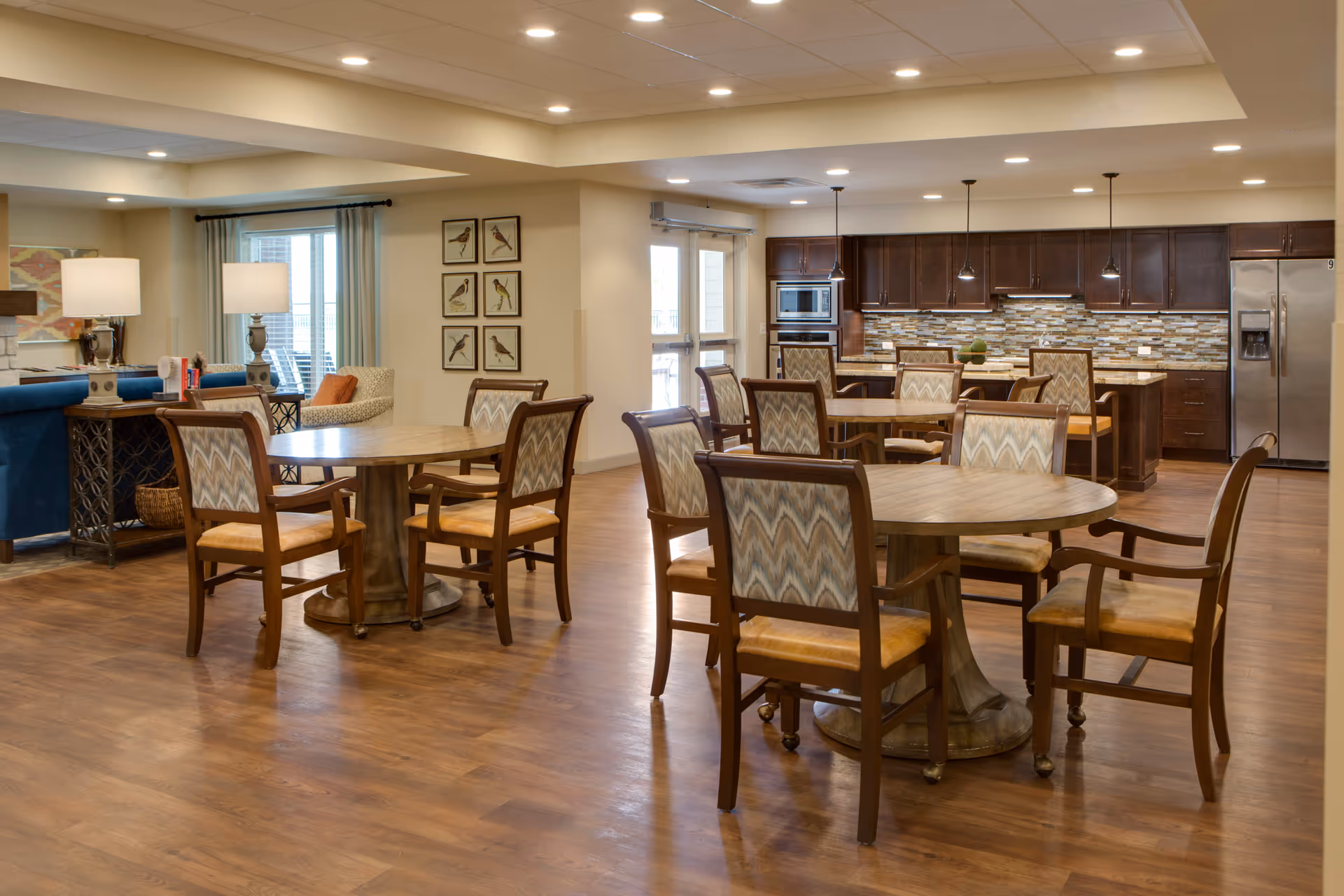 A spacious, well-lit common area featuring multiple round wooden tables with cushioned chairs arranged around them. In the background, there is a modern kitchen with dark wood cabinets, stainless steel appliances, and a tiled backsplash. The room has wood flooring, recessed lighting, and large windows with curtains allowing natural light to enter. There are also two table lamps on side tables near a blue sofa and an armchair, with framed bird artwork on the wall.