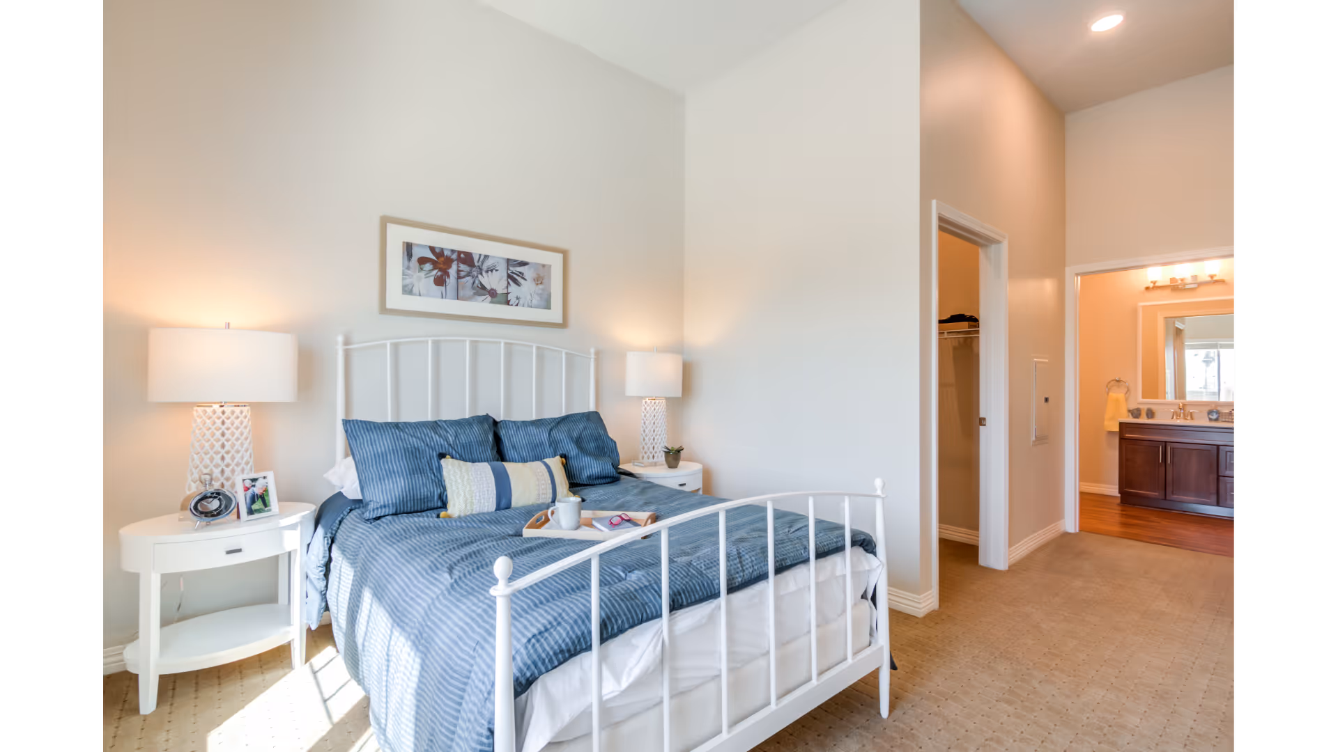 Bright bedroom featuring a white metal bed with blue bedding, matching nightstands and lamps, and an open doorway leading to a bathroom.