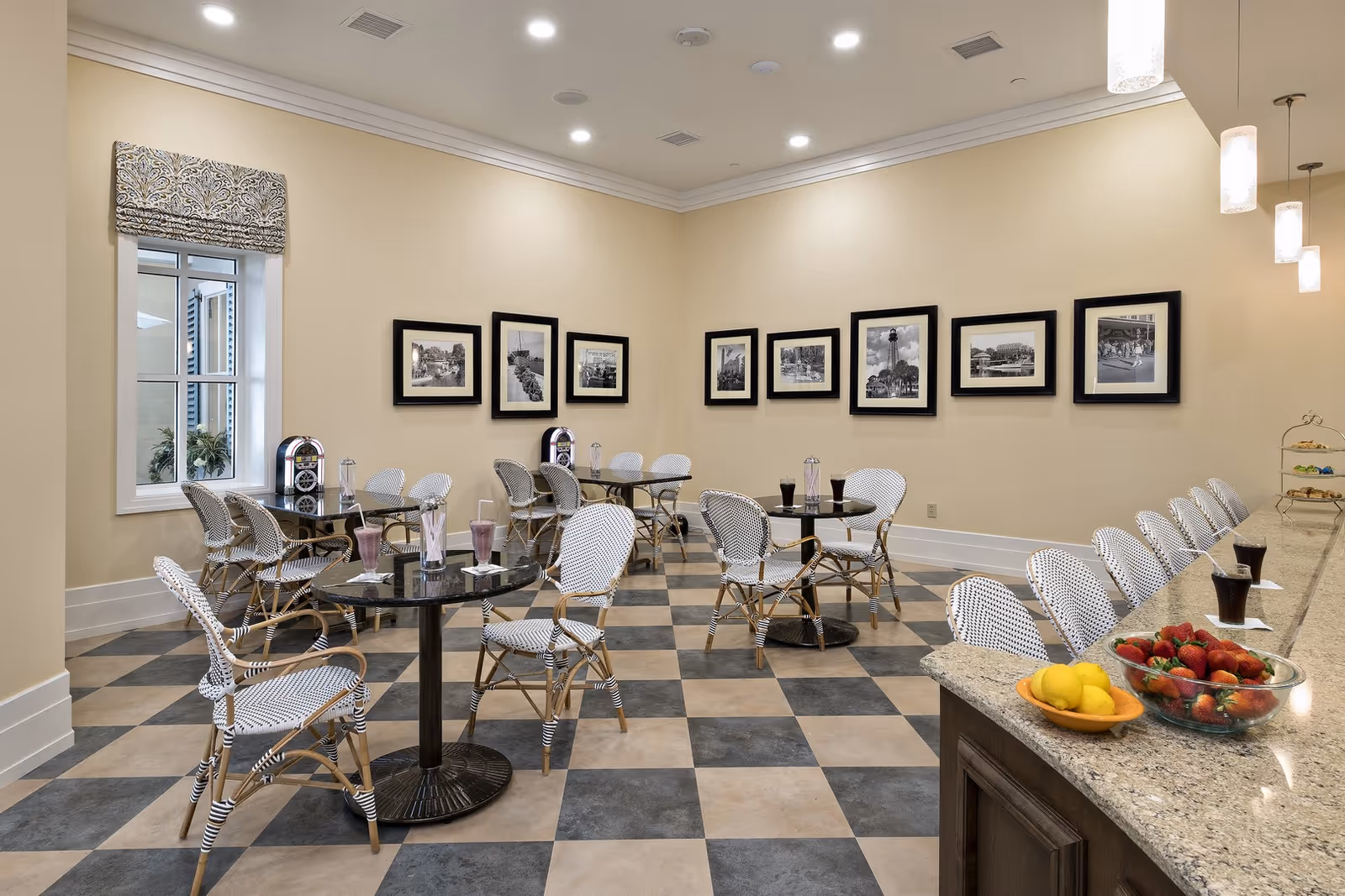 A bright dining area with checkered tile flooring, several round tables with wicker chairs, and a long granite countertop with high chairs. The tables have drinks and napkins on them, and the countertop has bowls of fruit including strawberries and lemons. The walls are decorated with framed black and white photographs, and there is a window with a patterned valance.