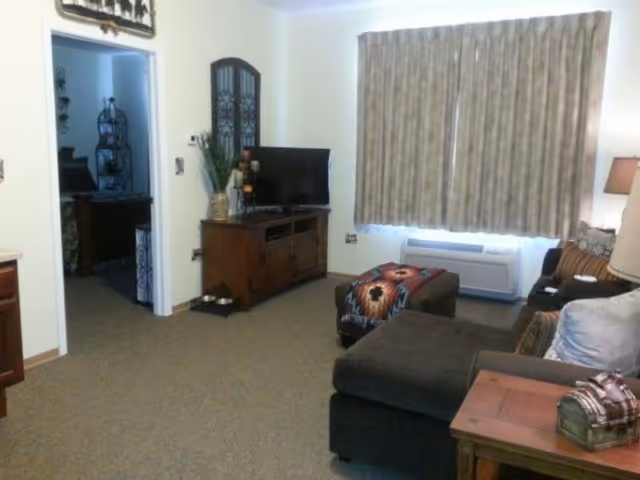A cozy living room with a dark sectional sofa, a wooden side table with a decorative chest, a TV on a wooden stand, and a window with closed beige curtains. There is a doorway leading to another room with visible furniture and decor.