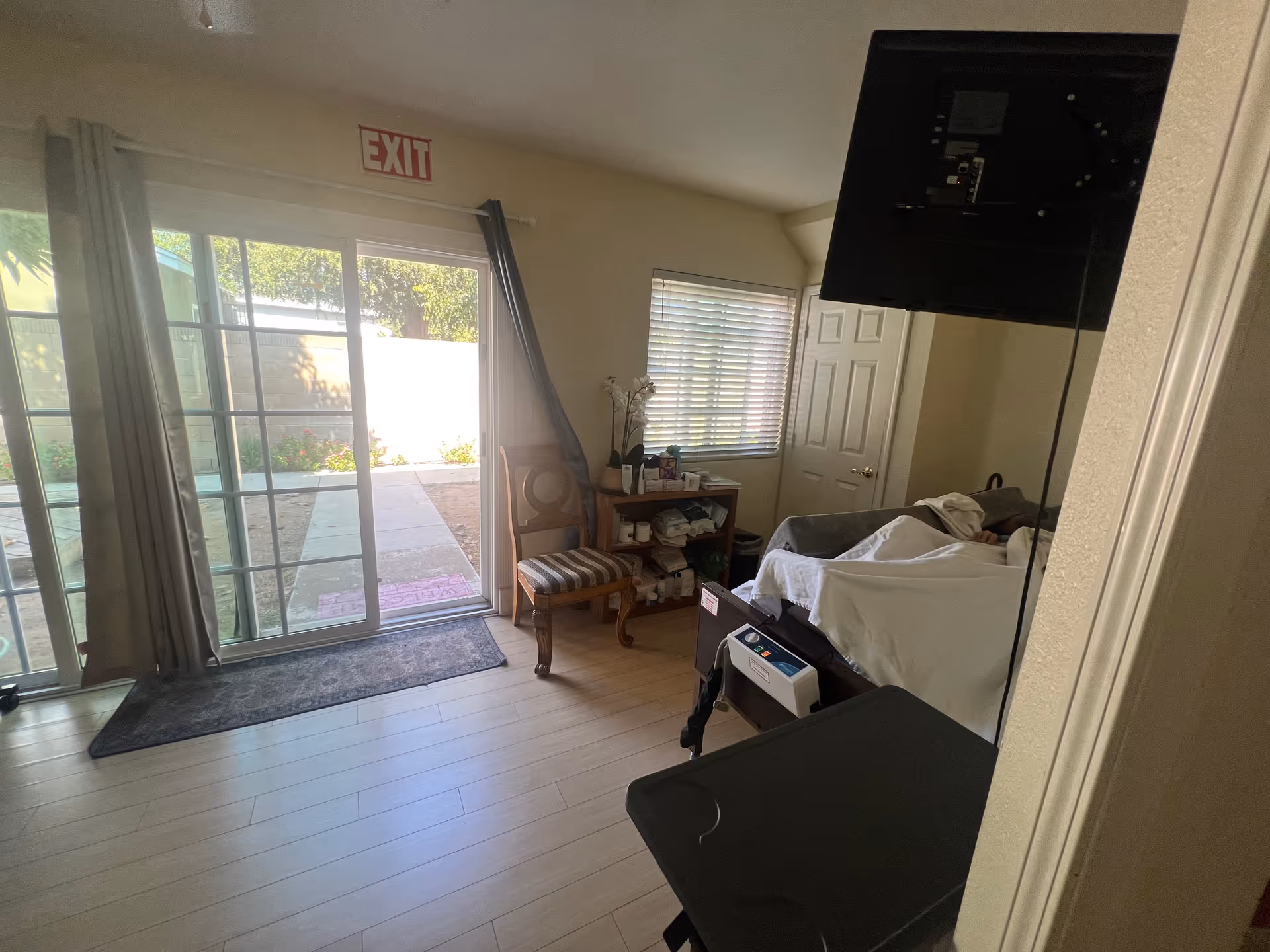 A small room with a hospital bed occupied by a person covered with a white blanket. The room has light wooden flooring, a wooden chair with a striped cushion, a small wooden shelf with various items, and a window with blinds. There is a sliding glass door with curtains leading to an outdoor area, and an exit sign above the door. A TV is mounted on the wall opposite the bed.