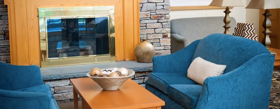 A cozy living room area featuring a blue upholstered loveseat and chair, a wooden coffee table with a decorative bowl of natural elements, a stone fireplace with a brass frame, and a beige sofa with a patterned pillow in the background.