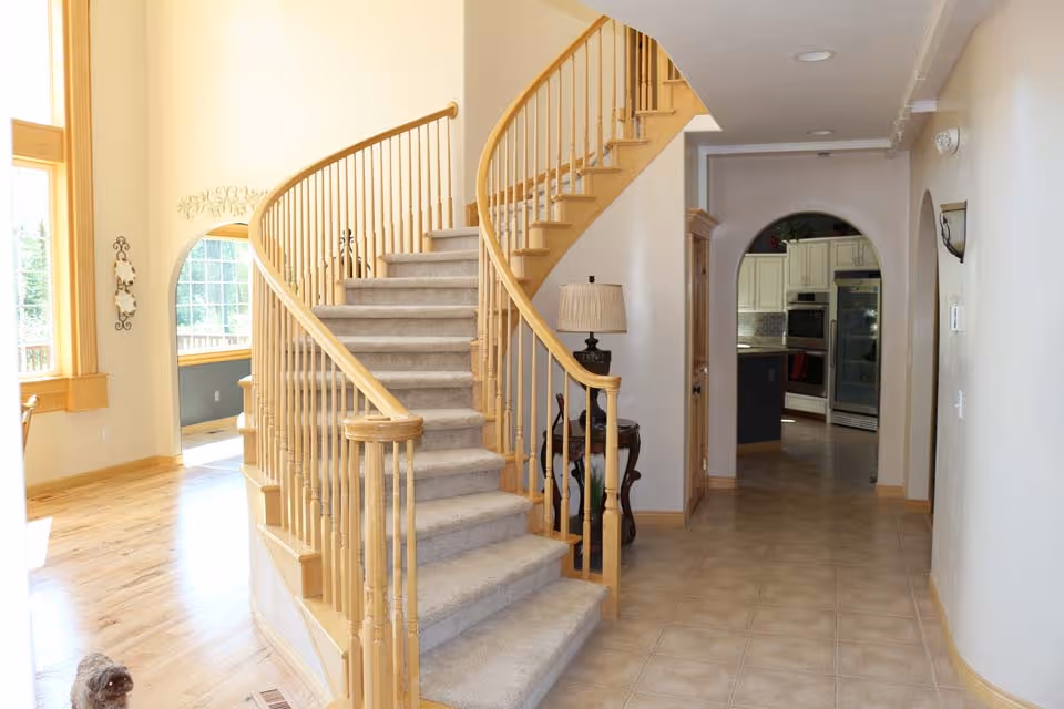 Interior view of a senior living facility showing a carpeted curved staircase with wooden railings. To the right, there is a hallway with tiled flooring leading to a kitchen area with white cabinets and stainless steel appliances. A small table with a lamp is positioned near the staircase. Large windows on the left side allow natural light to brighten the space.