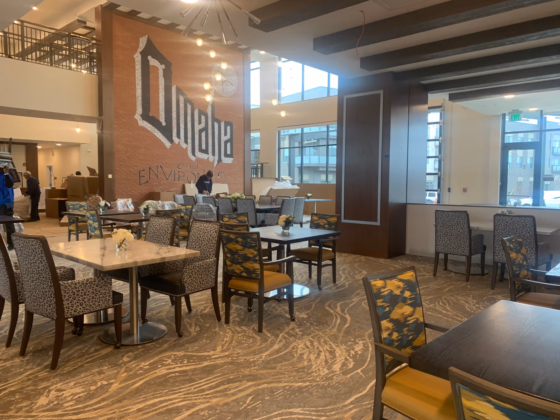 A spacious dining area with multiple tables and chairs, some with patterned upholstery. The room has large windows letting in natural light and a tall brick wall with the word 'Omaha' painted on it. There are decorative hanging lights and a carpeted floor with a swirling pattern.