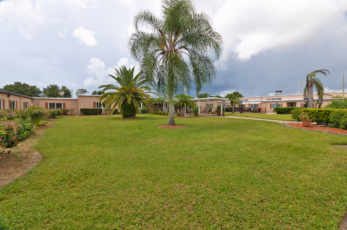 Well-kept grassy courtyard with palm trees and a single-story brick senior living building in the background under a cloudy sky.