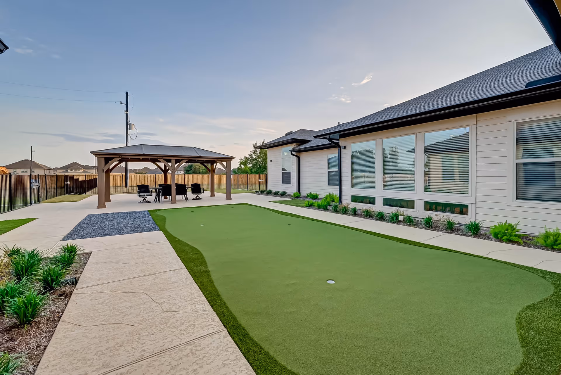 Outdoor area of a senior living facility featuring a putting green, a paved walkway, landscaped plants, and a covered seating area with chairs and a table. The building has large windows with blinds and a fenced yard in the background under a clear sky.