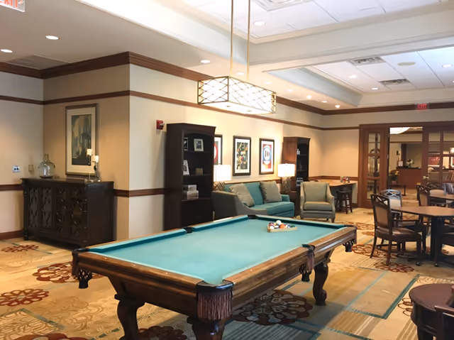 A spacious common area in a senior living facility featuring a pool table in the foreground, a seating area with a sofa and armchair against the wall, framed artwork above the seating, bookshelves, and several round tables with chairs. The room has warm lighting, patterned carpet, and a ceiling light fixture above the pool table.