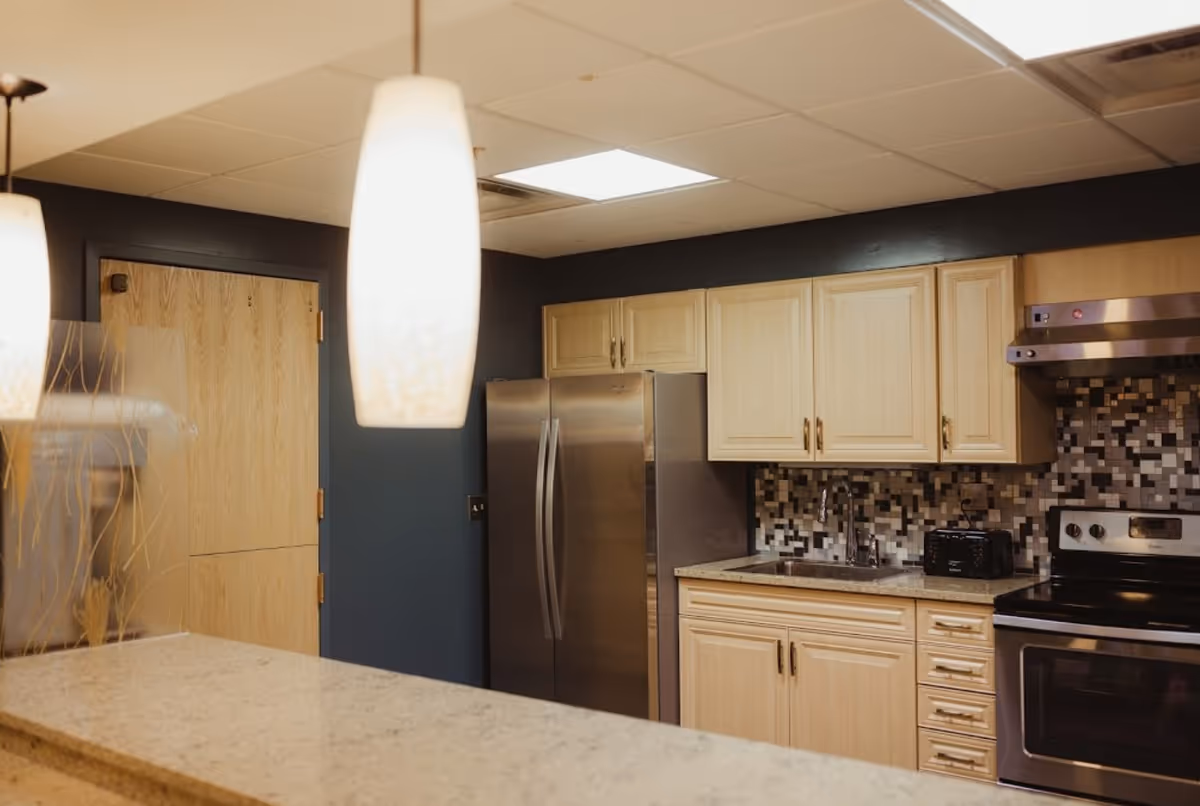 Modern kitchen with light wood cabinets, a stainless steel refrigerator, stove, and a mosaic tile backsplash. There is a granite countertop in the foreground and two pendant lights hanging from the ceiling.